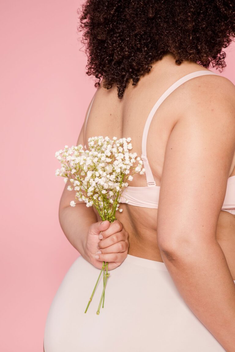 Back view of crop anonymous African American female in underwear with bouquet of blooming flowers against pink background