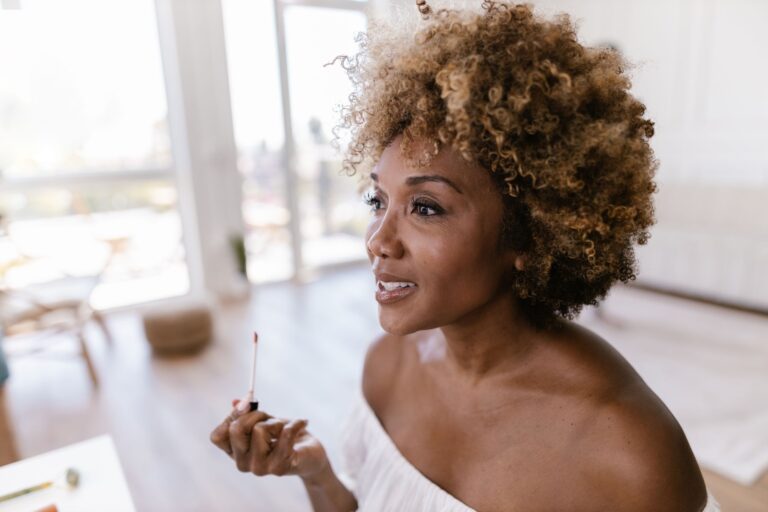 Confident woman with curly hair applying lipstick in a sunlit room, embracing natural beauty.